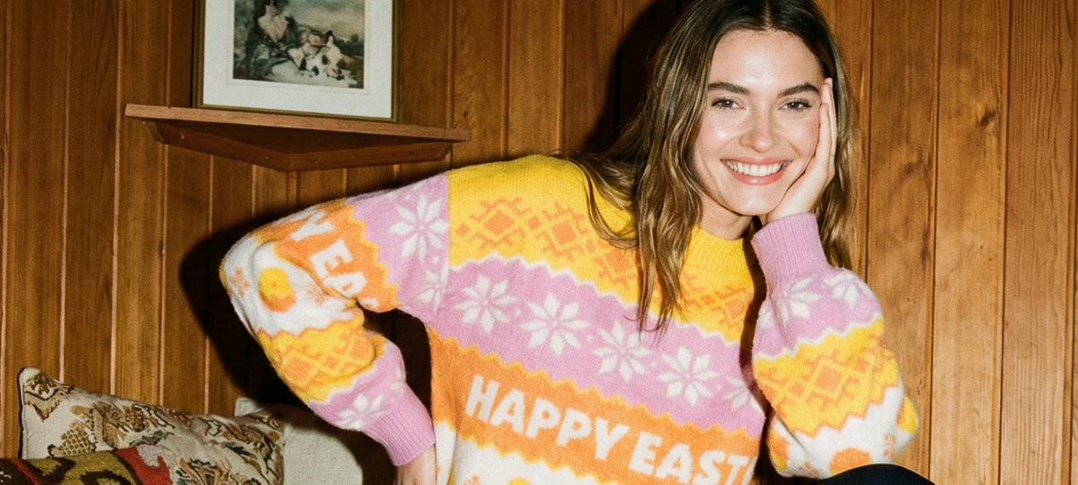 Smilende kvinne i fargerik strikkegenser med Happy Easter-mønster foran trepanelvegg